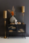 Black cabinet with decorative items against a dark wall, flanked by two gold floor lamps.