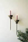 Two wall-mounted candle holders with pink candles on a white wall, accompanied by greenery.