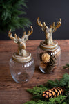 Two decorative glass jars with ribbed clear bodies and metallic gold deer head lids, displayed on a rustic wooden surface with evergreen branches and pinecones.