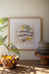 Framed artwork of fish and lemons on a wall with a plant and stack of plates below.