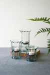 Three glass candle holders with candles on a gray surface, with a fern in the background.