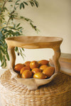 Wooden bowl with oranges on a woven surface with a plant in the background