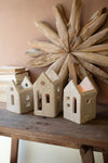 Three ceramic house-shaped candle holders on a wooden surface with a decorative wooden circle in the background.