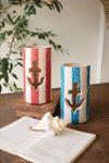 Two decorative candles with anchor designs on a wooden surface with a shell and paper.