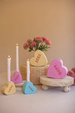 Decorative heart-shaped candles and tokens on a wooden stand with a vase of pink flowers in the background.