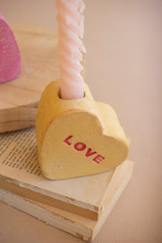 Heart-shaped object with 'LOVE' text on a stack of books, with a pink candle in the background.