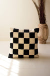 Black and white checkered pillow on a light surface with a wooden vase in the background.