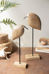 Two wooden bird sculptures on stands with a natural setting