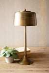 Brass table lamp on a wooden surface with a plant and books in the background