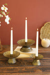 Three brass candle holders with white candles on a wooden surface against a red wall.