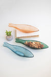 Two glass leaf-shaped platters, one blue and one green, on a white surface with a small plant in the background.