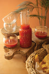 Tomato-based sauce in a blender with glasses on a wooden stand, surrounded by a plant and basket of chips.