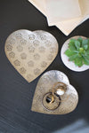 Heart-shaped metal object with floral patterns on a dark surface with a plant and paper in the background.