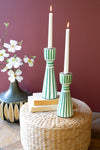 Green and white striped candle holders with candles on a woven ottoman against a purple wall.