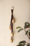 Decorative brass bells on a brown ribbon against a beige wall with greenery.