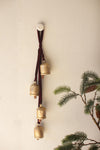 Brass bells on a leather strap against a neutral wall with greenery.