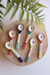 Colorful ceramic spoons with heart designs on a wooden board with a leafy branch in the background.