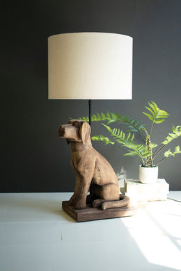 Wooden dog-shaped lamp with a beige lampshade on a white surface against a dark wall.