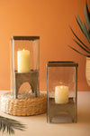 Two candle lanterns with candles inside on a neutral surface against an orange wall.