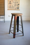 Vintage-style metal stool with wooden seat in a room with a poster on the wall.
