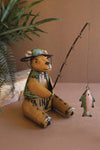 Teddy bear figurine dressed as a fisherman with a fishing rod and fish on a brown surface.