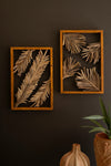 Two wall art pieces with leaf patterns in wooden frames on a dark wall.