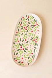 An oval ceramic serving platter with a festive holly and berry pattern, displayed on a warm beige background.