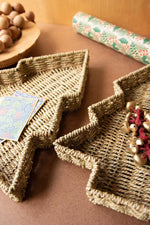 Two woven seagrass Christmas tree-shaped trays on a brown surface, styled with holiday decorations including a small card, gold jingle bells
