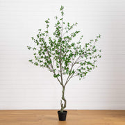 An artificial potted tree with green leaves and a twisted trunk, displayed in a black pot on a light wood floor against a white paneled wall.
