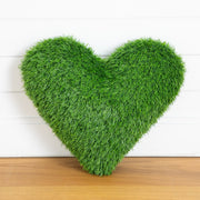 A heart-shaped decorative throw pillow made of realistic green grass or artificial turf, standing upright on a wooden surface against a white paneled wall.