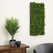 A rectangular artificial green foliage wall hanging mounted on a white wall, shown in a styled interior with a potted palm on a wooden sideboard and part of a gray chair visible.