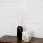 A black ceramic vase filled with delicate white decorative branches or twigs, styled on a dark wooden table next to two wrapped gift boxes against a white shiplap wall.