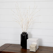 A black ceramic vase filled with delicate white decorative branches or twigs, styled on a dark wooden table next to two wrapped gift boxes against a white shiplap wall.