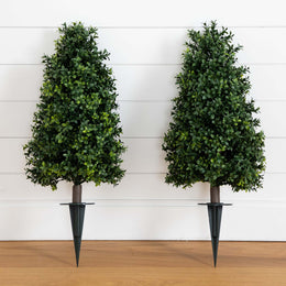 Two artificial boxwood topiary trees with dark metal spike planters, displayed side-by-side against a white shiplap wall and light wood floor.