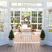 Sunroom with wooden floor, white walls, and large windows, featuring a wicker chair and potted plants.