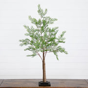 An artificial cypress tree with green foliage and a brown trunk, displayed on a black square base over a wooden table against a white shiplap wall.