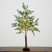 A small artificial Christmas tree with green pine-like foliage, warm white string lights, a brown trunk, and a black square base, displayed on a wooden surface against a white paneled wall.