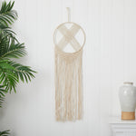Macrame wall hanging with fringes on a white wall with a plant and vase in the background