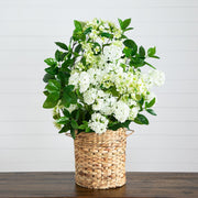 A faux floral arrangement of white flowers and green leaves in a woven natural fiber basket, displayed on a dark wooden surface against a white paneled wall.