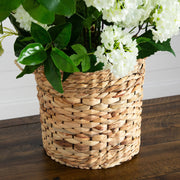 A natural woven seagrass basket planter holding white hydrangea flowers and green leaves, placed on a dark wood table against a white paneled wall.