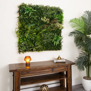 A square artificial green wall garden with various faux plants and succulents mounted on a white wall above a dark wood console table. A lit amber table lamp and a decorative brass object sit on the table, with a large potted palm plant visible on the right.