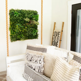 A styled interior scene showing a white sofa with cream and gray patterned pillows, a woven basket, and a green living wall hanging on the wall. A wooden ladder sign reading 'LOVE YOU MORE' stands nearby.
