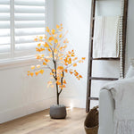 Artificial tree with orange leaves in a pot on a wooden floor next to a white wall with a window and a decorative ladder.