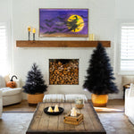 A styled living room featuring a large rectangular wall art print with a purple sky, black bats, and a witch riding a broomstick in front of a yellow full moon. The print is displayed on a wooden mantel above a fireplace with stacked firewood, flanked by two black feathered artificial trees in woven planters.