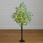 An artificial tree with green leaves and warm white string lights, standing on a dark trunk with a square base against a white paneled wall and wood floor.