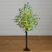 An artificial tree with green leaves and warm white string lights, standing on a dark trunk with a square base against a white paneled wall and wood floor.