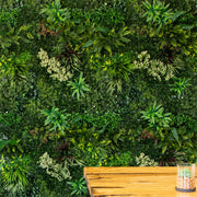 A lush artificial green wall mural featuring various faux ferns, moss, and small plants, with a wooden tabletop and a small glass planter visible in the bottom right corner.