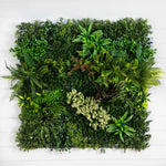 A square artificial living green wall made of dense faux foliage and assorted plants, mounted against a white wood plank background.