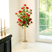 An artificial potted camellia tree with vibrant red flowers and green leaves, standing in a white ceramic pot on a tiled floor next to a window.