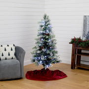 A flocked artificial Christmas tree with white lights and pinecones stands on a red velvet tree skirt in a living room corner with a gray armchair and wooden side table.